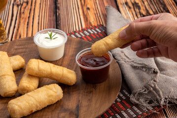 typical Venezuelan fried cheese tequeños on a round wooden plate with two sauces and hand dipping the tequeño in red sauce