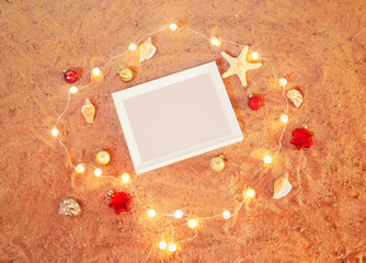 White frame, chrismas decorations, glowing christmas lights, starfishes and seashells on sand beach. Top view with copy space.