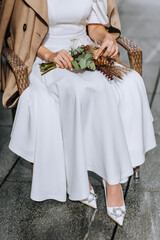 A beautiful bride in a white dress, beige coat sits in an armchair with a bouquet of wild flowers in her hands with a golden ring on her finger outdoors. Wedding autumn photography.