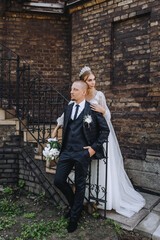 A young groom in a blue plaid suit and a lovely bride in a white lace dress with a diadem and a bouquet are embracing, standing near the wall on the stairs of the church. wedding photography.