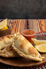 Argentinian meat or chicken empanadas on a round wooden plate with lemon wedges and bowls of hot...