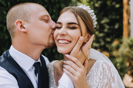 Wedding photo of cheerful, smiling newlyweds. A stylish, young groom and a beautiful bride in a white dress with a bouquet in their hands, a diadem on their heads, sit in a park in nature.