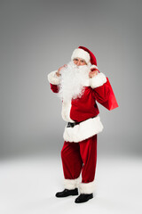 Full length of father christmas in costume holding shopping bag while standing on grey background.