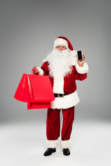 Santa claus in costume holding shopping bags and smartphone with blank screen on grey background.