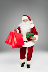 Full length of santa claus holding red shopping bags and gift on grey background.