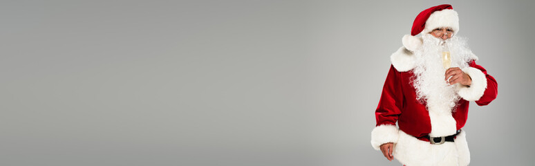Santa claus in red costume and eyeglasses holding glass of champagne isolated on grey, banner.
