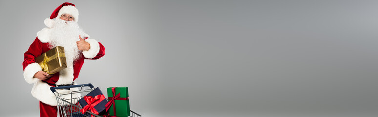 Father christmas showing thumb up near shopping cart with presents isolated on grey, banner.