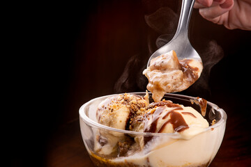 vanilla ice cream cup dessert with brownie and hot fudge syrup with spoon in female hand tasting...