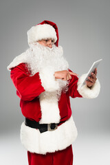 Santa claus in eyeglasses pointing at digital tablet isolated on grey.