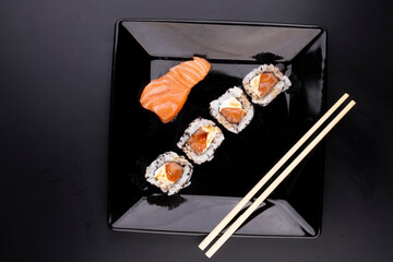 a piece of raw salmon sashimi japanese food on black plate with 4 more salmon and cream cheese sushis with soy sauce and chopsticks in top view