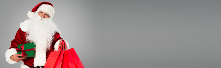 Bearded santa claus in costume holding shopping bags and present isolated on grey, banner.