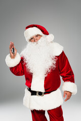 Santa claus in red costume holding car key and looking at camera isolated on grey.