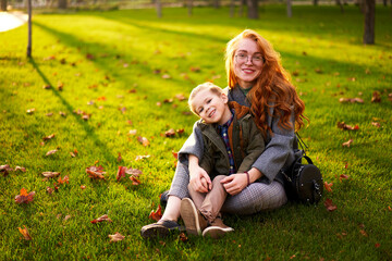 Naklejka na ściany i meble Smiling redhead woman and little boy sitting on grass lawn in city park on warm autumn day. Young mom hugs her son, they have fun and look at camera on sunny fall day. Foliage on green lawn.
