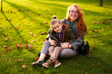 Smiling redhead woman and little boy sitting on grass lawn in city park on warm autumn day. Young mom hugs her son, they have fun and look at camera on sunny fall day. Foliage on green lawn.