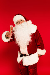 Santa claus in eyeglasses and costume holding champagne glass on red background.