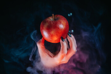 Woman as witch offers red apple as symbol of temptation, poison. Fairy tale, white snow wizard concept. Spooky halloween, cosplay. Smoke, haze background.