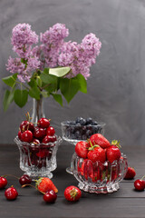 On the table in small glass vases are strawberries, cherries, blueberries, a bouquet of lilacs on the background. Open space