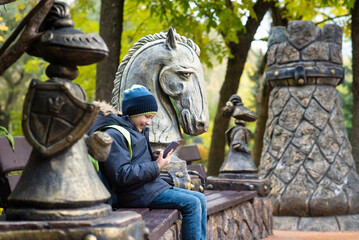 Fototapeta na wymiar a boy sits on a bench among sculptures in the form of chess pieces in the park, plays online chess on his phone. in Presnensky park