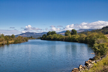 On a sunny Autumn day in Northwestern Idaho along the Trail of the Coeur d'Alenes, the partly cloudy blue sky is reflected in the surface of a peaceful river with mountains in the distance.