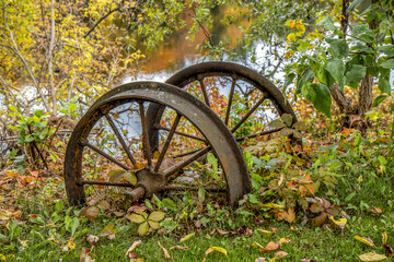 Set of rusted steel wagon wheels with a wooden axle, sitting outdoors in grass and autumn leaves on a river bank, nobody