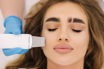 Close up of female face with soft skin. Woman at cosmetology clinic taking a beauty procedure for skin. Beautician using peeling device, ultrasonic clining. Gorgeous woman - obrazy, fototapety, plakaty