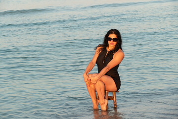 The girl is sitting on a stool in the sea. Brunette in a black swimsuit on the beach.