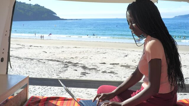 Young digital nomad woman working from her camper van. Woman remote worker using her laptop inside a motorhome in front of a beautiful beach