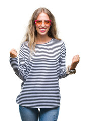 Beautiful young blonde woman wearing sunglasses over isolated background celebrating surprised and amazed for success with arms raised and open eyes. Winner concept.