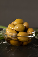 Olives in an extra virgin olive oil placed in a glass bowl and green branches on a dark table with...