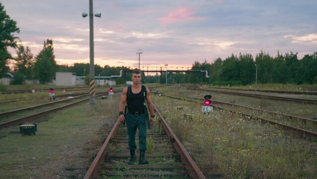 Young muscular man in sleeveless T-shirt goes between the rails against the backdrop of pipeline and semaphores glowing red. Slow motion