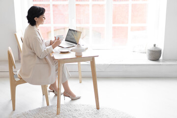 Woman sits at a desk near a window in a spacious light interior. Coffee break. Copy space.