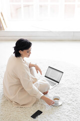 Female milf with black hair in a cream suit sits on a cozy carpet on the floor. Coffee break. Working on a laptop in a minimalist home living room.