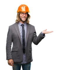 Young handsome architec man with long hair wearing safety helmet over isolated background smiling cheerful presenting and pointing with palm of hand looking at the camera.