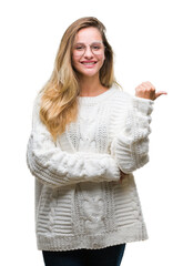 Young beautiful blonde woman wearing winter sweater and sunglasses over isolated background smiling with happy face looking and pointing to the side with thumb up.