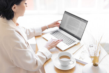 Woman in a jacket is typing in a text document on her laptop. Sitting at the desk with a cup of...