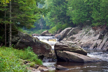 Misty  river waterfall flowing through large boulders in upstate new york, adirondack region, surrounded by large trees and dense forest.