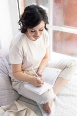 An adult satisfied woman with false eyelashes sits on a wide windowsill. Makes entries in his diary.