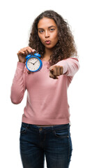 Young hispanic woman holding alarm clock pointing with finger to the camera and to you, hand sign, positive and confident gesture from the front