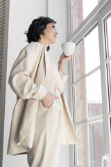 Woman in a business beige suit drinks coffee in office standing in front of the window. Coffee...