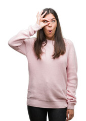 Young beautiful hispanic woman wearing a sweater doing ok gesture shocked with surprised face, eye looking through fingers. Unbelieving expression.