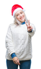 Young blonde woman wearing christmas hat over isolated background smiling with happy face winking at the camera doing victory sign. Number two.