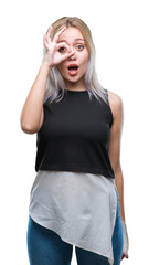 Young blonde woman over isolated background doing ok gesture shocked with surprised face, eye looking through fingers. Unbelieving expression.