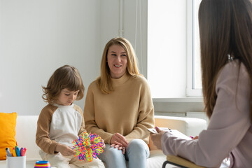 Young cheerful mother at a reception at a child family psychologist with her little son. Little boy with long haircut plays educational toys. Blurred foreground. - obrazy, fototapety, plakaty
