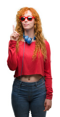 Young redhead woman wearing headphones surprised with an idea or question pointing finger with happy face, number one