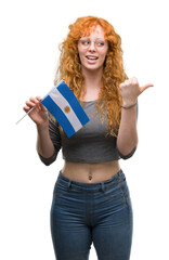 Young redhead woman holding flag of Argentina pointing and showing with thumb up to the side with happy face smiling