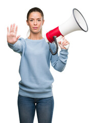 Beautiful young woman holding megaphone with open hand doing stop sign with serious and confident expression, defense gesture