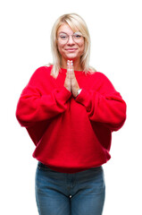 Young beautiful blonde woman wearing sweater and glasses over isolated background praying with hands together asking for forgiveness smiling confident.