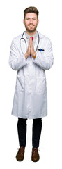 Young handsome doctor man wearing medical coat praying with hands together asking for forgiveness smiling confident.