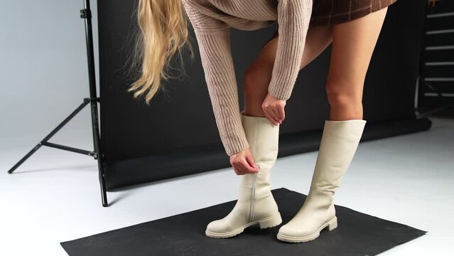Blond long haired lady bends to check her elegant light high boot. Model footage of stylish modern footwear on plain soles and low heel.