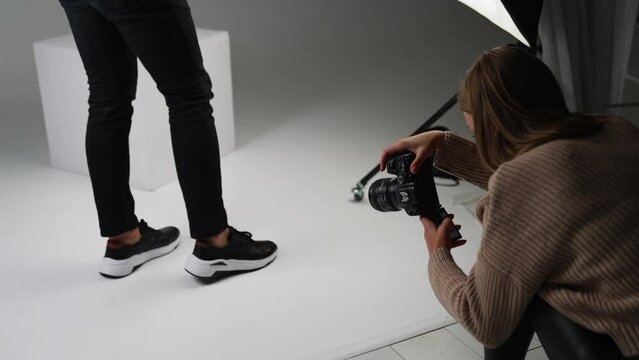 Lady taking shot of male feet in new modern black sneakers with a white sole. Backstage work of a photographer in studio.